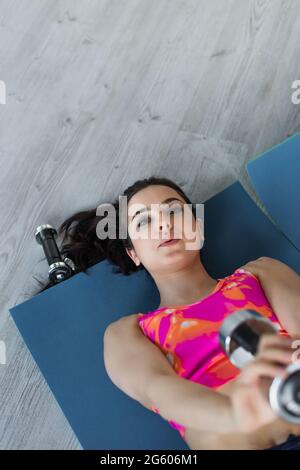 top view of focused young woman in sportswear lying on fitness mats and doing exercise with dumbbells at home Stock Photo