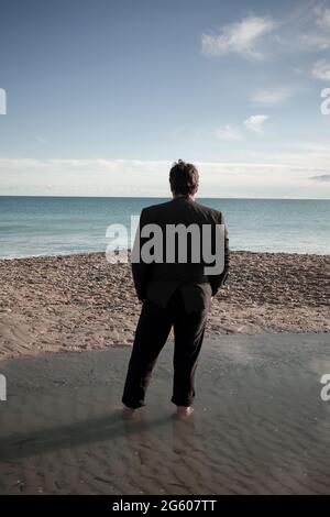 rear view of tall man with dark hair in dark suit with his hands in pockets, standing in beach puddle looking out across a calm sea Stock Photo