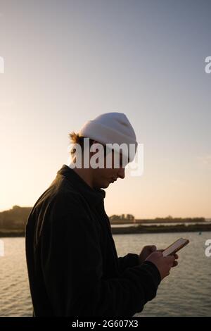 side profile of twenty year old man with cream beanie hat standing by a canal checking messages on his smartphone during an early morning walk Stock Photo