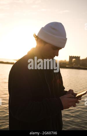 side profile of twenty year old man with cream beanie hat standing by a canal checking messages on his smartphone during an early morning walk Stock Photo