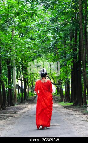 Young woman wearing a red saree walking on green asphalt road. The girl walks along the path in the woods to the light. Stock Photo