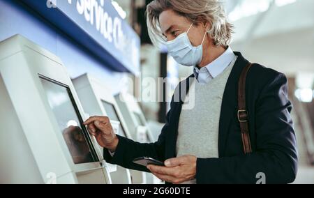 Business traveler using self service check in at airport. Man on business trip wearing face mask doing self checking during pandemic. Stock Photo