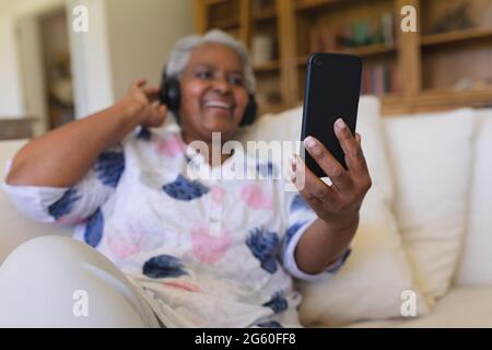 Senior african american woman sitting on sofa wearing headphones using smartphone Stock Photo