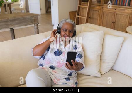 Senior african american woman sitting on sofa wearing headphones using smartphone Stock Photo