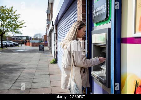 A side-view shot of a mid-adult caucasian woman using a debit card at a cash point on a city street, she's wearing casual clothing. Stock Photo