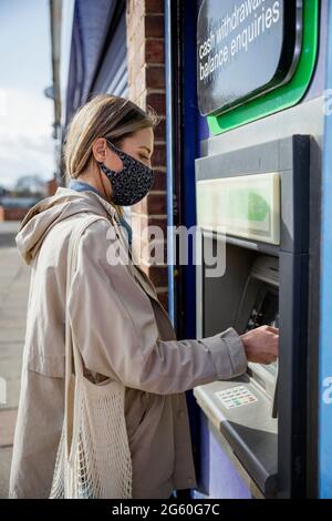 A side-view shot of a mid-adult caucasian woman using a debit card at a cash point on a city street, she's wearing casual clothing and a protective fa Stock Photo