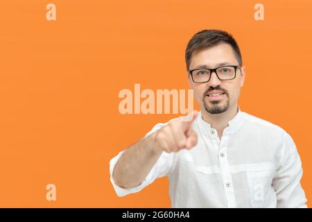 Cheerful caucasian man with beard wearing white shirt and glasses pointing to you and camera. Handsome smiling positive man. Orange background with co Stock Photo