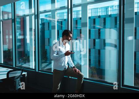 Mixed race businessman at window using smartphone and looking away Stock Photo