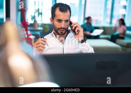 Mixed race businessman sitting at table using smartphone with colleagues in background Stock Photo