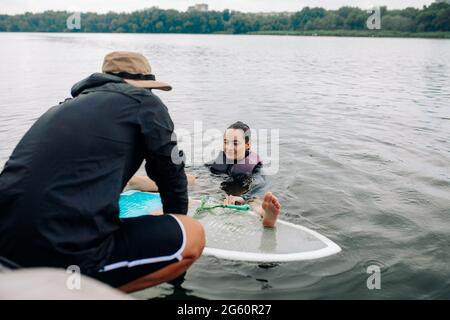 Instructor teaches young woman wakesurfing technique and she learns how to stand up on surf board while holding to tow rope. Stock Photo