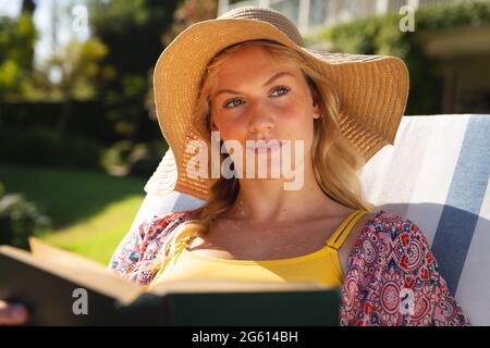 Happy caucasian woman wearing sunhat relaxing in sunny garden reading book, looking away, smiling Stock Photo