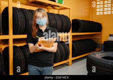 portrait of asian female worker wear a mask to prevent the spread of corona virus or COVID-19 checking the stock of car tires at warehouse Stock Photo