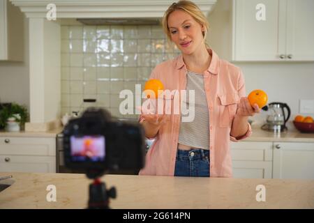 Smiling caucasian woman in kitchen holding oranges and using camera, making cooking vlog Stock Photo