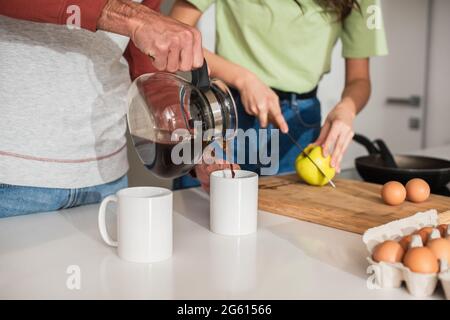 Cropped view of man pouring coffee near blurred girlfriend cutting apple in kitchen Stock Photo