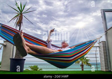Businessman doing a video conference with the smartphone lying in a hammock in a terrace close to the sea. In a cloudy day with the sun beams illumina Stock Photo