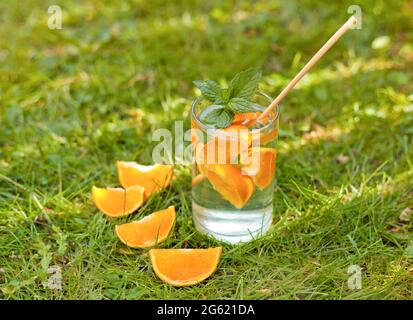 cold lemonade drink with orange and mint in tall glass on green grass and eco straw. glass with oranges and water on the grass Stock Photo