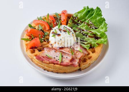 Hearty Belgian waffles with poached egg, bacon, lettuce, tomato and arugula, garnished with sesame seeds and green onions. Dish on a white plate. Stock Photo