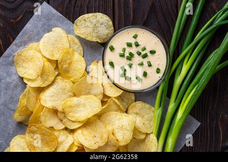 Crunchy potato chips, bunch of fresh green onions and mayonnaise on a black wooden table. Snack of deep-fried thin potato slices. Unhealthy eating. Stock Photo