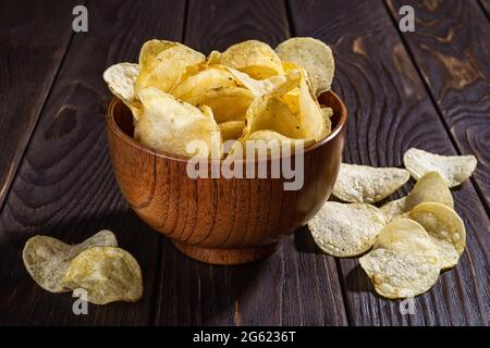 Crispy organic potato chips with dill in a brown wooden bowl on a black wooden table. Crunchy snack of deep-fried potato slices. Fast food, junk food. Stock Photo