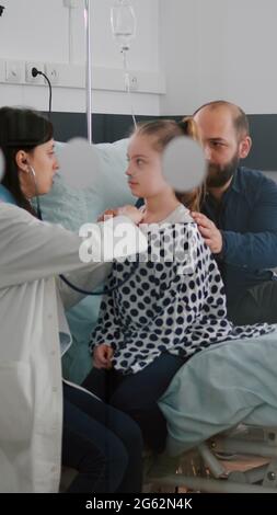 Paediatric doctor examining chest listening heartbeat using medical stethoscope while black woman nurse writing disease treatment working in hospital ward. Kid patient recovery after surgery Stock Photo