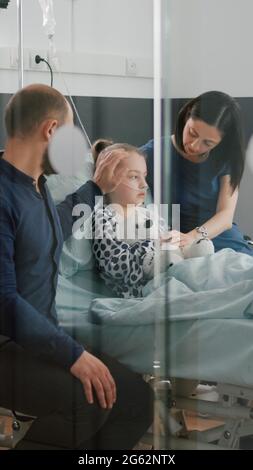 Sick daughter wearing oxygen nasal tube lying in bed after illness surgery waiting for medical examination with parents in hospital ward. Hospitalized child recovering after flu isickness nfection Stock Photo