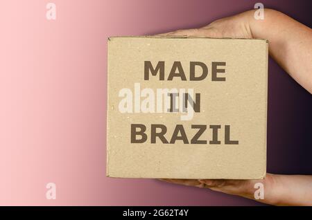 Hands holding cardboard box of Brazilian merchandise on colored gradient background. Man with a square package of recycled material. Inscription made Stock Photo