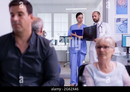 Specialist doctor holding patient x-ray explaining disease diagnosis to nurse while standing in hospital waiting area. Disabled senior woman in wheelchair waiting for medical examination Stock Photo