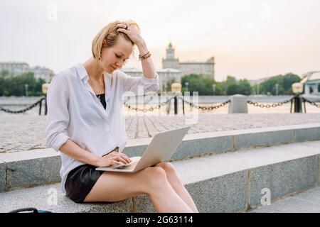 Young woman dressed in office clothes works or studies on a city street on her laptop.  Stock Photo