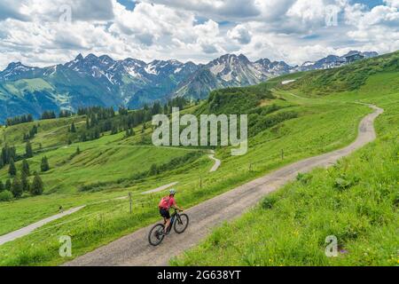 nice senior woman on mountain bike climbing up Mount Fellhorn in the Allgaeu High Alps with Trettach and Maedelegabel in background, Allgau, Bavaria, Stock Photo