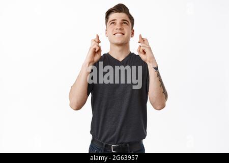 Wishful thinking. Hopeful smiling young man, student biting lip and looking up while making wish, holds fingers crossed for good luck, praying and Stock Photo