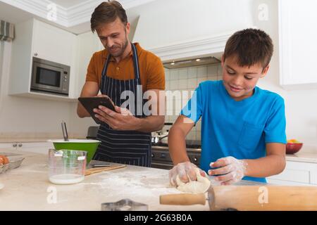 Caucasian father using tablet and son baking and smiling in kitchen Stock Photo