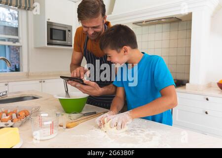Caucasian father using tablet and son baking and smiling in kitchen Stock Photo