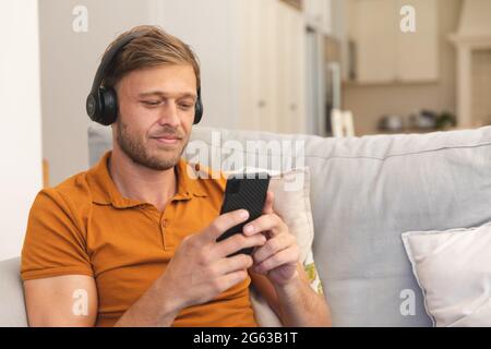 Caucasian man sitting on couch, wearing headphones and using smartphone at home Stock Photo