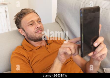 Caucasian man lying on couch, smiling and using tablet at home Stock Photo