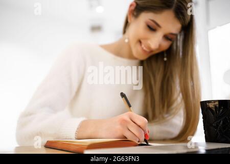 Smiling beautiful student female with long hair in white sweater writing in notebook while using the laptop. Remote Studing Stock Photo