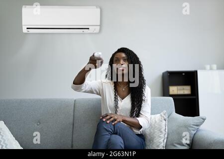Young Happy Woman Sitting On Couch Operating Air Conditioner With Remote Control At Home Stock Photo