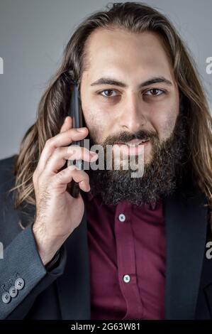 portrait of a young middle eastern businessman with beard and long hair while having a conversation on the mobile phone Stock Photo