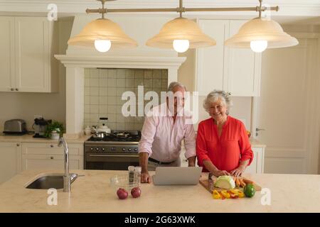 Portrait of senior caucasian couple cooking together and using laptop in kitchen Stock Photo