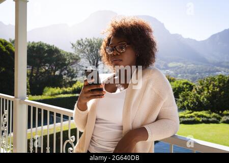Happy african american woman standing on sunny balcony of country home using smartphone Stock Photo