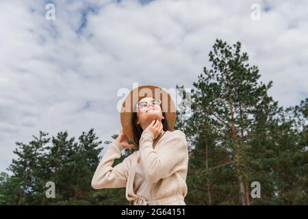 low angle view of cheerful young woman in sunglasses and straw hat looking up near forest Stock Photo