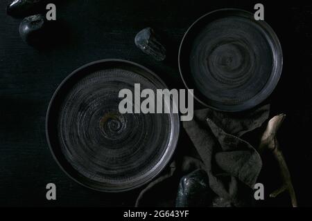 Set of empty black ceramic plates with black stones and wooden around on textile napkin over black wooden background. Flat lay. Stock Photo