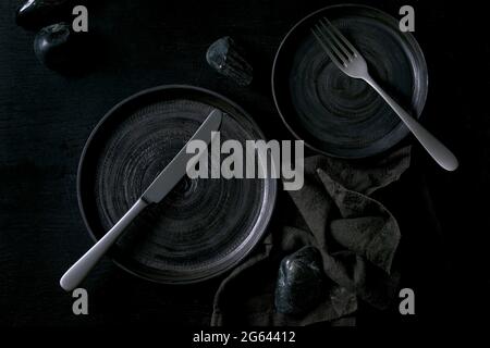 Empty black ceramic plates with cutlery and black stones around on textile napkin over black wooden background. Flat lay, space. Stock Photo