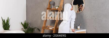 young woman in bathrobe with towel on head brushing teeth in bathroom near mirror, banner Stock Photo
