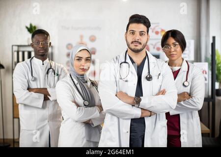 Team of multiracial doctors in white lab coats keeping hands crossed while standing together at medical center. Concept of people, medicine and teamwork. Stock Photo