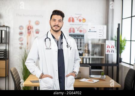 Portrait of confident indian doctor keeping hands in pockets of white lab coat while posing at hospital room. Concept of people, health care and treatment. Stock Photo