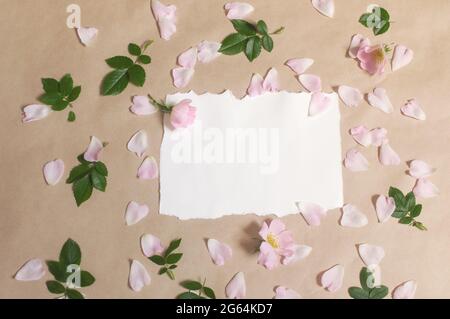 blank paper, pink buds flowers and rose petals, green leaves are spread out on a beige background Stock Photo