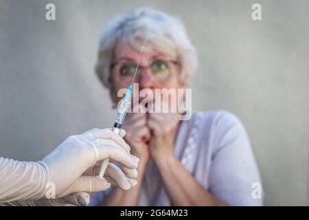 Hands in medical gloves are holding a syringe, preparing for vaccination, in the background an elderly woman in glasses is afraid to give an injection Stock Photo