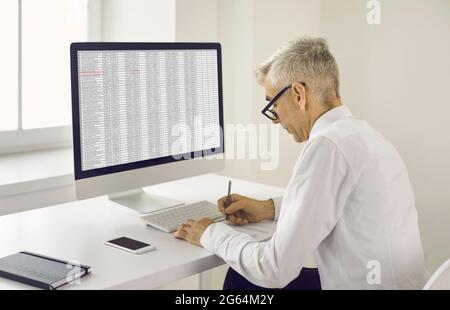 Senior man taking notes on paper while sitting at office desk with desktop computer Stock Photo