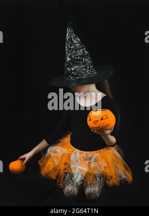 A girl in a witch costume - a hat with a spider web, an orange skirt-holds a pumpkin bucket for collecting sweets in her hands Stock Photo