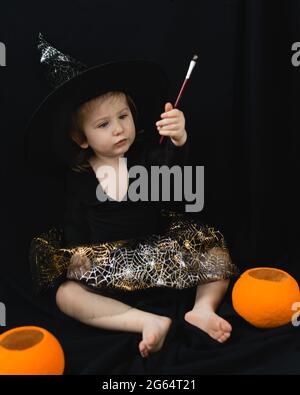 A little cheerful girl in a witch costume-a hat and a skirt with a spider web-sits on a black background and examines a magic wand. Next to it is a bu Stock Photo